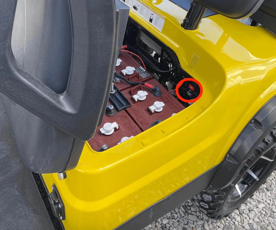 Open seat compartment of a yellow golf cart, with the switch next to the batteries circled